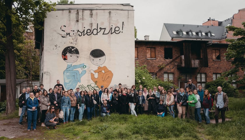 Uczestnicy spaceru sąsiedzkiego pozujący do grupowego zdjęcia w plenerze. W tle budynek z muralem. Zdjęcie archiwalne