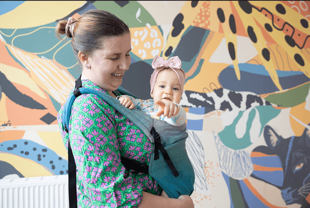 Mama Ela z małą Marysią w chuście; Marysia paluszkiem celuje w obiektyw fotoreportera
