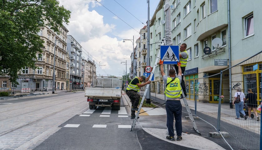 Trójka robotników w żółtych kamizelkach montuje znak drogowy przy przejściu dla pieszych i przejeździe dla rowerzystów / przebudowa Pomorskiej i Staszica / 28 maja 2024