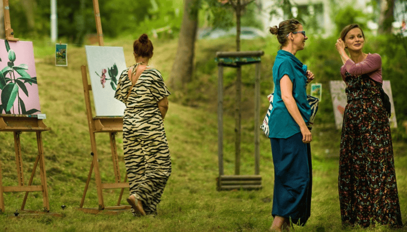 Trzy kobiety, po lewej jedna ubrana w długą suknię ogląda plenerową wystawę, po prawej stoją dwie kobiety  