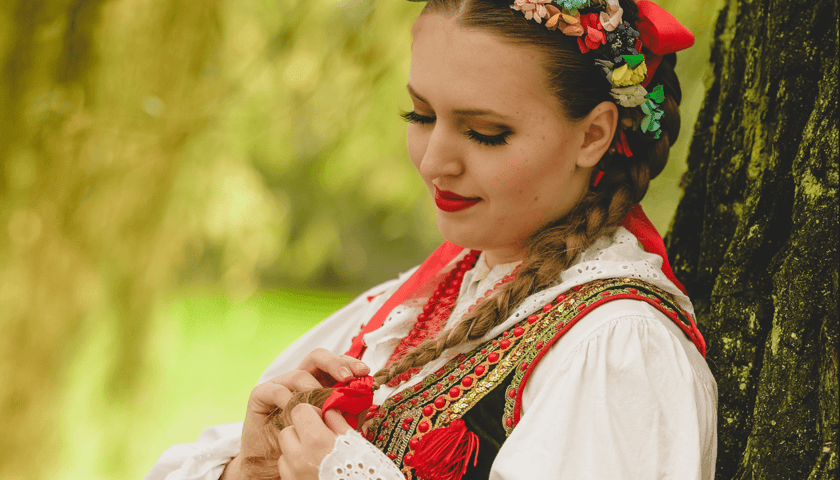 Dziewczyna w stroju ludowym stojąc przy drzewie ogląda kokardę na końcu swego warkocza - zdjęcie zrobione podczas sesji Zespołu Pieśni i Tańca KALINA w dworku w Ożarowie w roku 2023