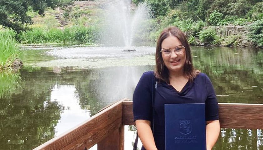 Uśmiechnięta kobieta w granatowej sukience, w dłoniach trzyma teczkę na dyplom. W tle zbiornik wodny z fontanną i zieleń. Zuzanna Rzemińska, absolwentka Wydziału Inżynierii Środowiska
