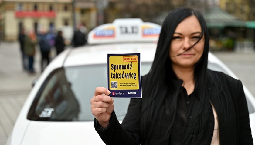 Kobieta trzyma w ręku ulotkę z napisem - sprawdź taksówkę, w tle widać taksówkę