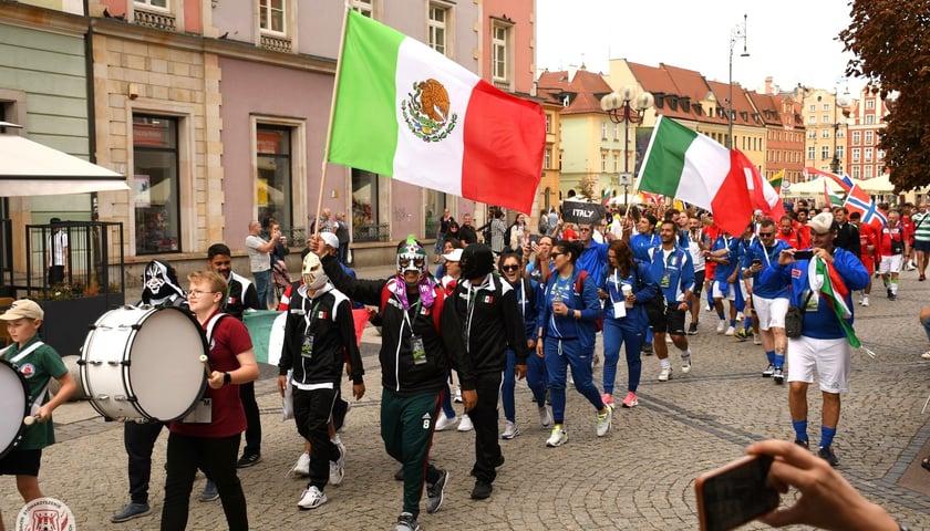 Tłum ludzi: na przodzie z bębnami, dalej w maskach i flagą Meksyku, niebieskich dresach z flagą Włoch. Pochód zawodników  Międzynarodowy Turniej Piłki Nożnej Ulicznej Osób Bezdomnych 2023