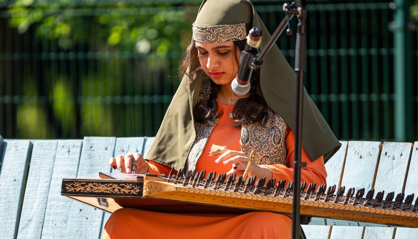 Dziewczynka w pomarańczowym ludowym stroju gra na instrumencie (podobnym do cytry)  