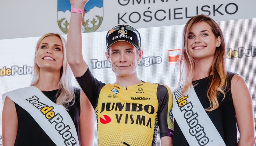 Uśmiechnięty Jonas Vingegaard (w środku) w żółtej koszulce, obok niego dwie kobiety z szarfami z napisem Tour de Pologne