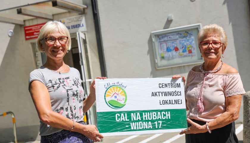 Wejście do CAL na Hubach przy ul. Widnej 17. Na zdjęciu wolontariuszki CAL na Hubach Ewa Fic i Teresa Sroczyńska trzymające tabliczkę z napisem "Centrum Aktywności Lokalnej CAL na Hubach - Widna 17"