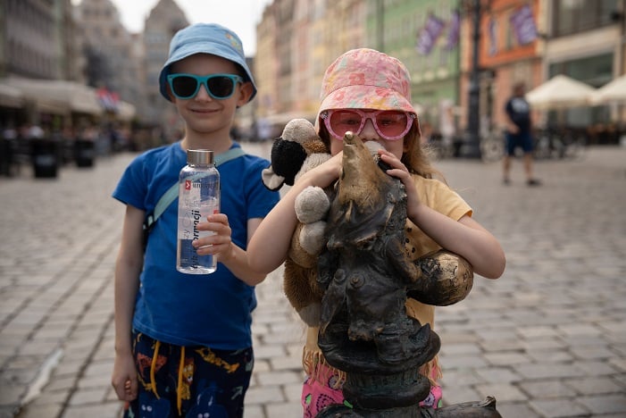 Chłopczyk i dziewczynka na wrocławskim Rynku: on trzyma w ręce bidon z wodą, ona opiera się o krasnala-zraszacza