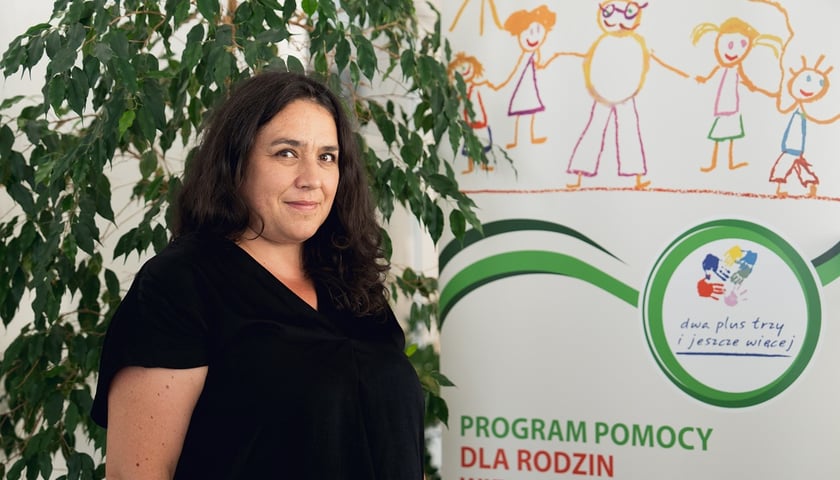 Anna Powietrzyńska z Miejskiego Ośrodka Pomocy Społecznej we Wrocławiu na tle plakatu programu Dwa plus trzy plus jeszcze więcej