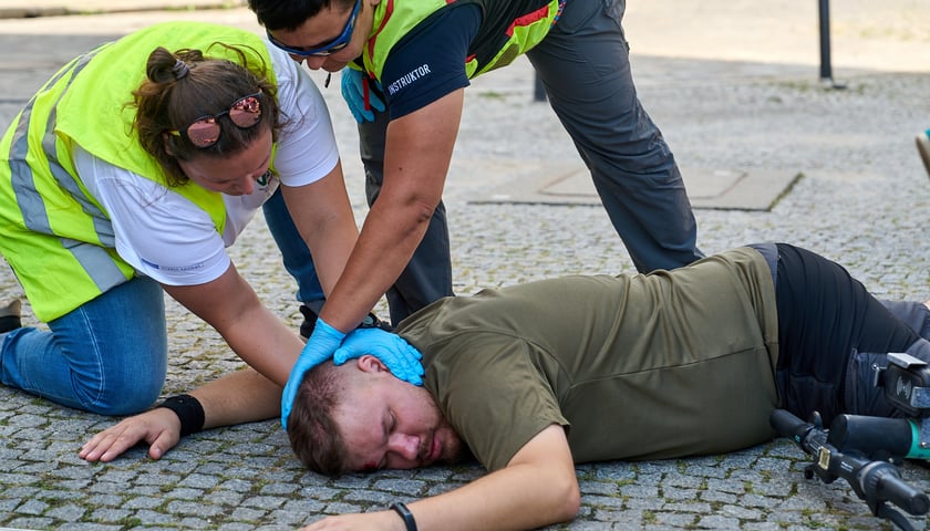 Na bruku leży mężczyzna w krótkich spodenkach, obok w odblaskowych kamizelkach klęczą dwie osoby i udzielają poszkodowanemu pierwszej pomocy