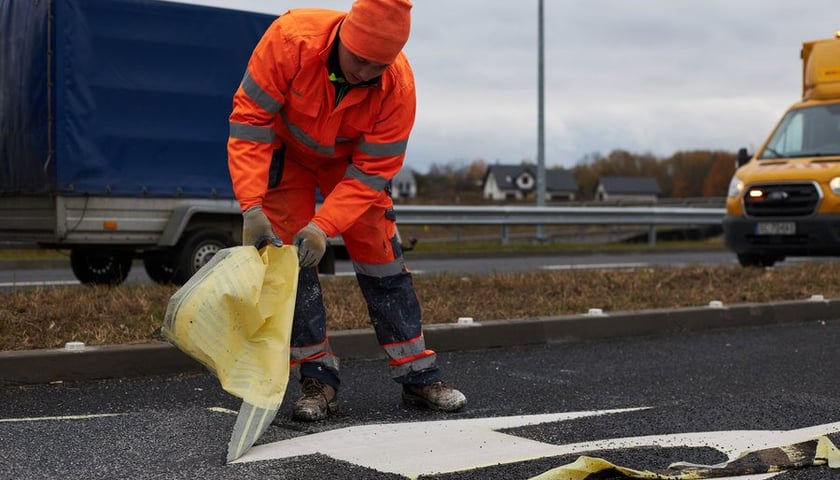 Mężczyzna w pomarańczowym stroju roboczym zrywający taśmy po malowaniu znaków poziomych na WOW (zdjęcie z grudnia 2022)
