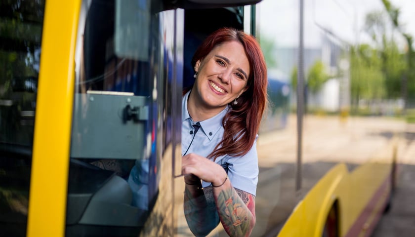 Za kierownicą autobusu siedzi uśmiechnięta kobieta z kolorowym tatuażem na  przedramieniu