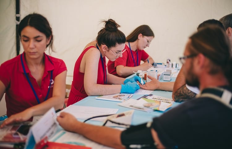 Trzy kobiety w ciemnoróżowych uniformach przy stole z osobami, którze przyszły w zeszłym roku na badania profilaktyczne