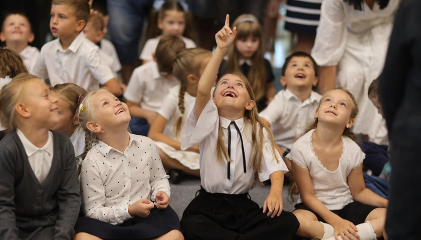 Dziewczynka w galowym ubraniu siedząc po turecku pokazuje palcem na coś w górze, na co patrzą z zachwytem również inne dzieci  - początek roku szkolnego w Zespole Szkolno-Przedszkolnym przy ul. Cynamonowej