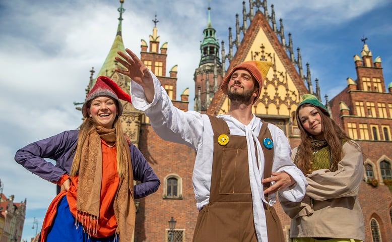 Trzy osoby - dwie kobiety i mężczyzna - przebrane za krasnale (spiczaste czapki, kolorowe ubrania) na tle wrocławskiego ratusza