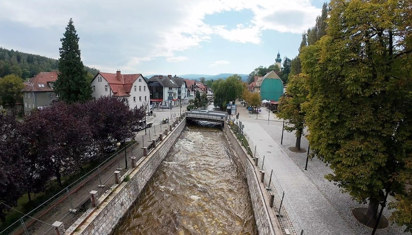 Widok na centrum Polanicy-Zdrój. Na środku zdjęcia widać rzekę, po boku miejsca spacerowe, zabudowania, drzewa...