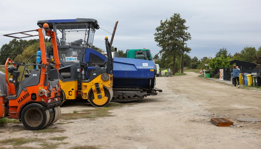 Maszyny budowlane na gruntowym kawałku ul. Lechitów, w tle widać budowlańców w żółtych kamizelkach