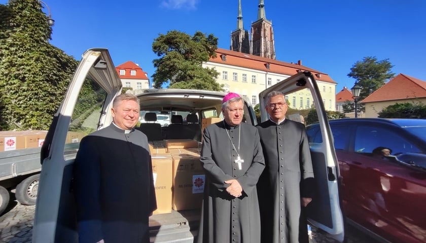 Przed busem z paczkami stoi trójka duchownych w sutannach, od lewej: ks. Dariusz Amrogowicz, dyrektor Caritas Archidiecezji Wrocławskiej, arcybiskup Jerzy Kupny i ks. proboszcz Krzysztof Dudojć z Lewina Brzeskiego