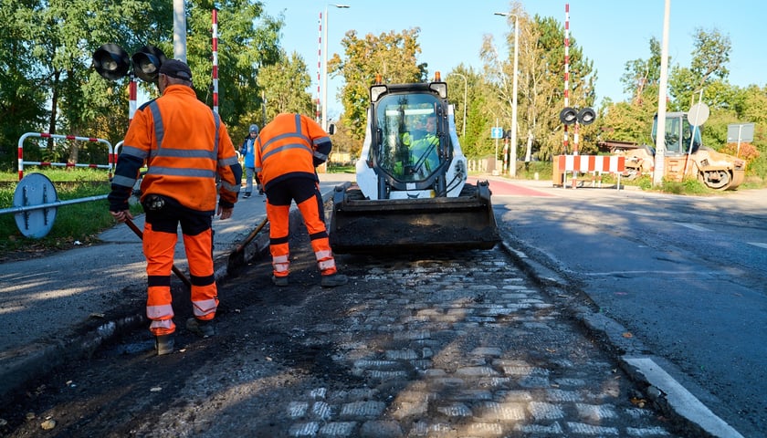 Dwaj mężczyźni w pomarańczowych kombinezonach pracują na ul. Ołtaszyńskiej, widać też maszynę budowalną 