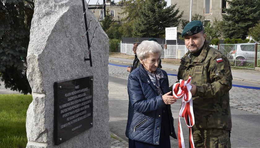 Kobieta w kurtce (siwe włosy) i żołnierz, trzymają biało-czerwoną wstęgę, przy pomniku na Leśnicy (24 października 2023)