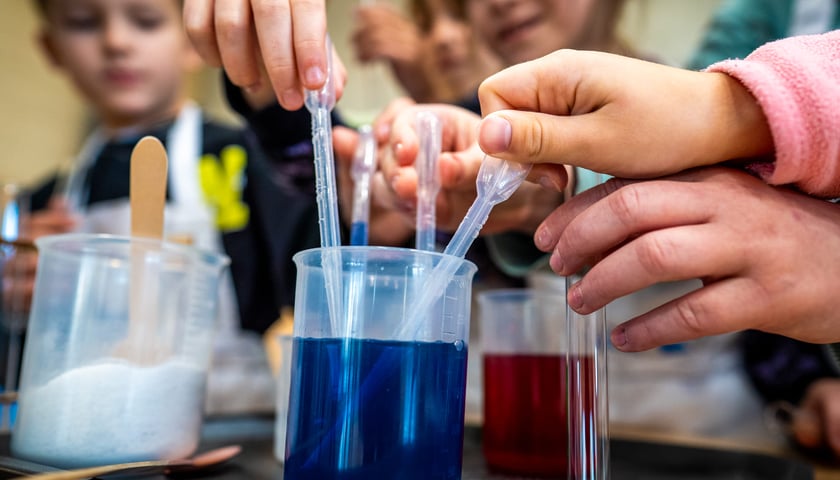 Akademia H2o z MPWiK Wrocław. Na zdjęciu naczynia z kolorowymi płynami. W tle rozmyte sylwetki dzieci biorących udział w chemicznym doświadczeniu.