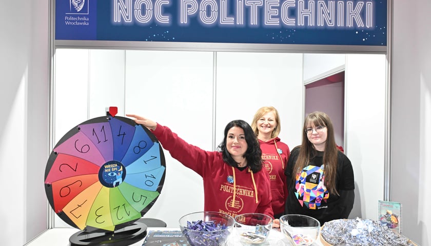 Trzy uśmiechnięte studentki Politechniki stoją przy Kole Fortuny. Powyżej napis "Noc Politechniki"