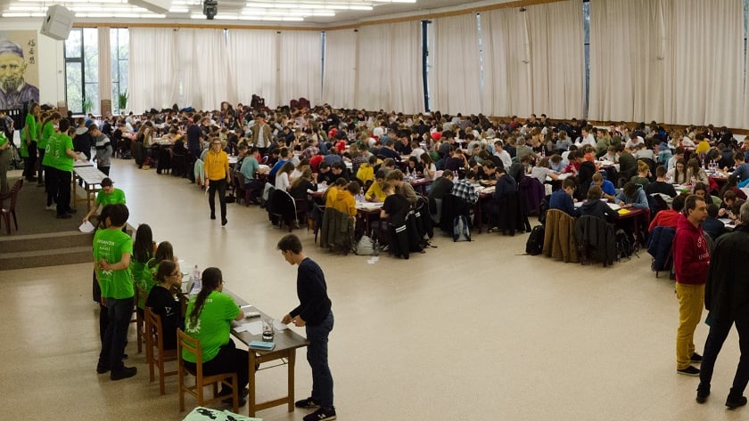 Duża sala wypełniona stolikami, przy którcy siedzą młodzi ludzie i rozwiązują zadania