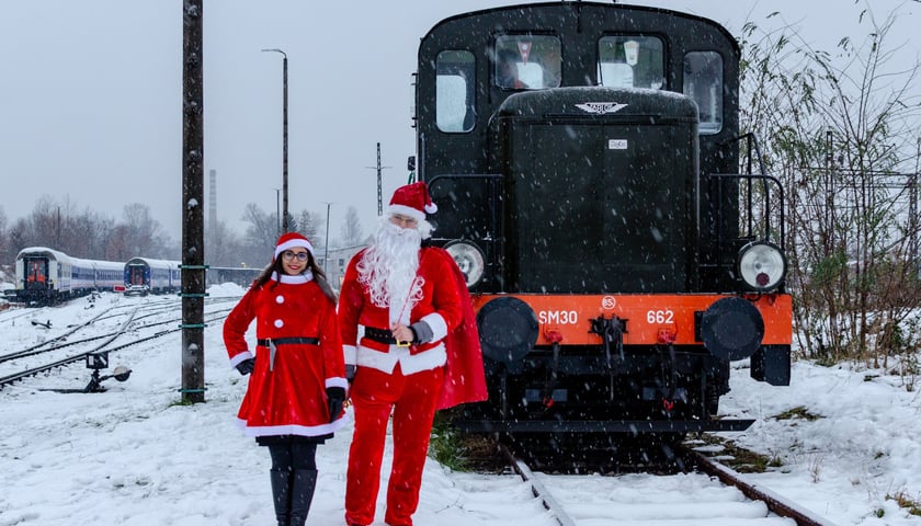 Śnieżynka i Święty Mikołaj (ubrani w czerwone, świąteczne stroje) stoją na tle pociągu retro 