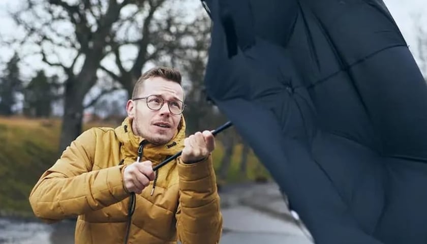 Młody mężczyzna w okularach trzyma w rękach parasol targany przez silne porywy wiatru