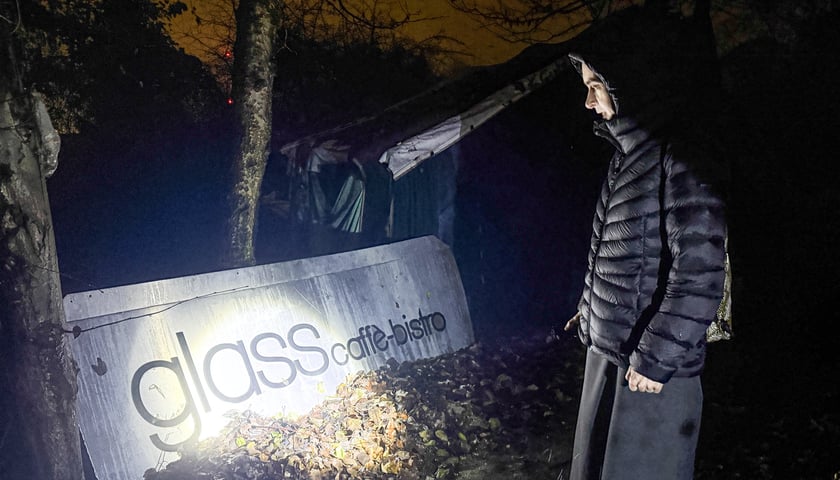 Kobieta, siostra zakonna z latarką, stoi przed szyldem nieistniejącego bistro z napisem Glass.