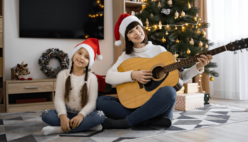 Dziewczynka i kobieta z gitarą siedzące przy choince, obie w czapkach czerwonych z białym pomponem