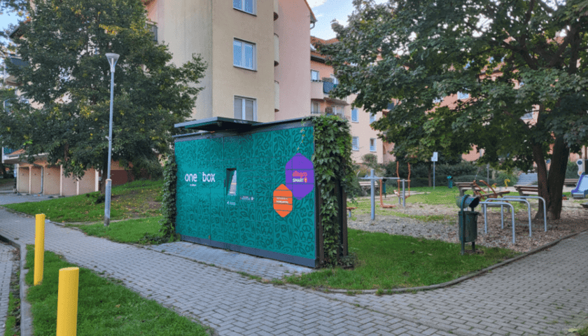 Automat kurierski oklejony zielonym motywem roślinnym, porośnięty rośliną pnącą, wzdłuż chodnika, na tle placu zabaw.
