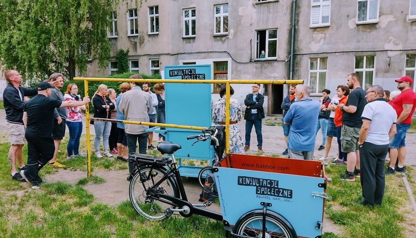 Rozmowa mieszkańców podwórka podczas konsultacji osiedlowych. Na pierwszym planie błękitny rower i wózek  konsultacyjny.