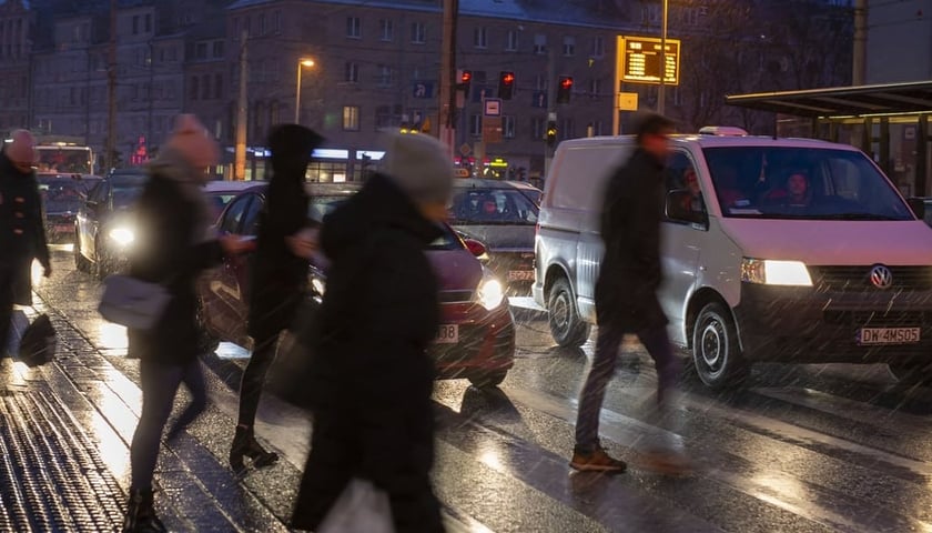 Przez ulicę przechodzą ludzie, widać także auta. Zdjęcie zrobione zimą, po zmroku