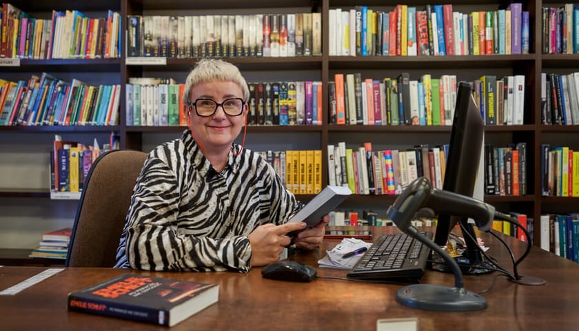Bibliotekarka Małgorzata siedzi przy biurku, trzyma książkę (filia biblioteki publicznej przy ul. Jesiennej). Zdjęcie ilustracyjne