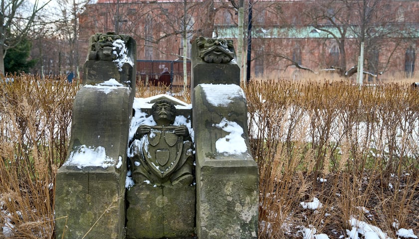Fragment kamieniarki z rzeźbą mężczyzny i dwiema innymi przedstawiającymi zwierzęta, na tle placu i budynku z czerwonej cegły
