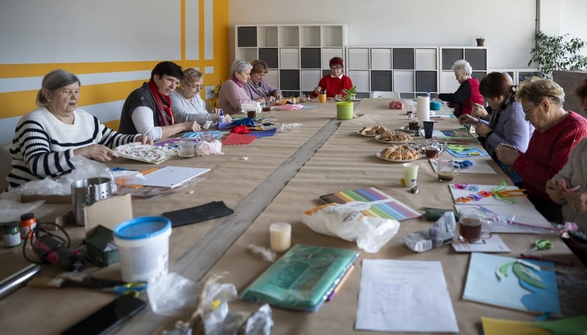 9 osób przy dużym stole maluje obrazy, na stole stoją materiały malarskie