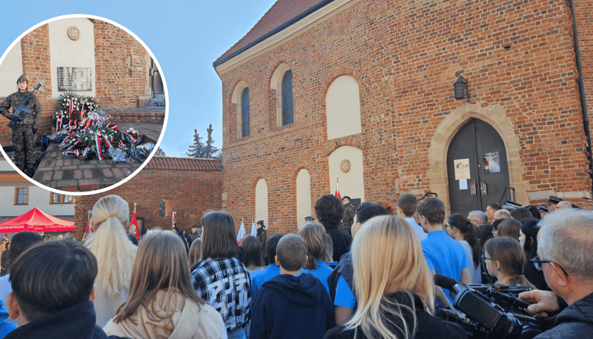 Tłum ludzi stojących tyłem do obiektywu, w kółku żołnierz i kwiaty pod tablicą na elewacji kościoła