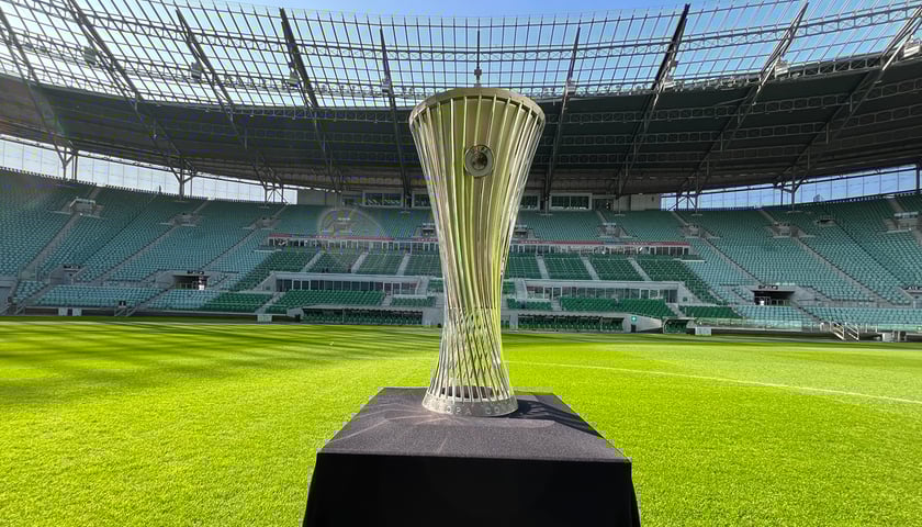 Na zdjęciu wysoki, lśniący puchar na postumencie na murawie stadionu