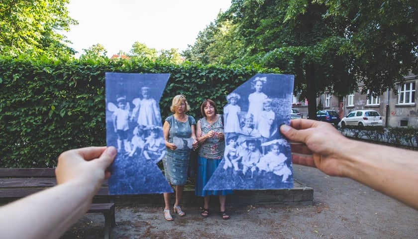 Przecięta fotografia z dawnych lat i dwie trzymające je ręce. W świetle przecięcia widać dwie kobiety stojące na tle żywopłotu (zdjęcie z realizacji projektu o historii Placu Grunwaldzkiego)