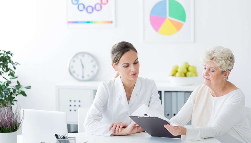 Lekarka w białym fartuchu siedzi przy biurku naprzeciwko starszej pacjentki i omawia z nią plan leczenia diabetologicznego. W tle widoczne są plakaty edukacyjne oraz miska z owocami.