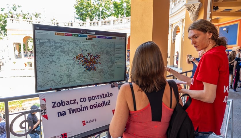 Kobieta i mężczyzna stoją przed monitorem, na którym wyświetlona jest baza #InwestycjeWRO
