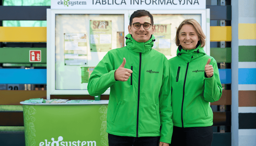 Mężczyzna i kobieta w zielonych kurtach z napisem Ekosystem na tle tablicy informacyjnej w nowym PSZOK-u przy ul. Szwajcarskiej