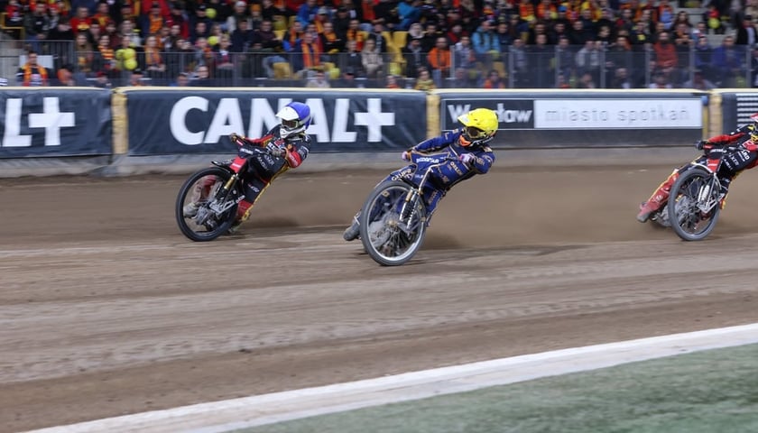 Trzech żużlowców walczy o zwycięstwo w jednym z wyścigów na stadionie Olimpijskim. W tle kibice w żółto-czerwonych szalikach.