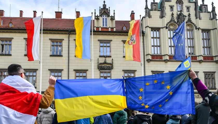 Flagi wiszące przed wrocławskim ratuszem: polska, ukraińska, wrocławska i Unii Europejskiej. Na pierwszym planie dwie osoby trzymają związane ze sobą rogami flagi Ukrainy i Unii Europejskiej. Zdjęcie archiwalne.