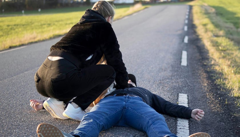Kobieta udzielająca na drodze pierwszej pomocy poszkodowanemu leżącemu na drodze.