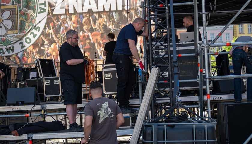 Trzech mężczyzn na budowanej scenie w Rynku, uchwyceni w momencie montowania jednego z jej elementów