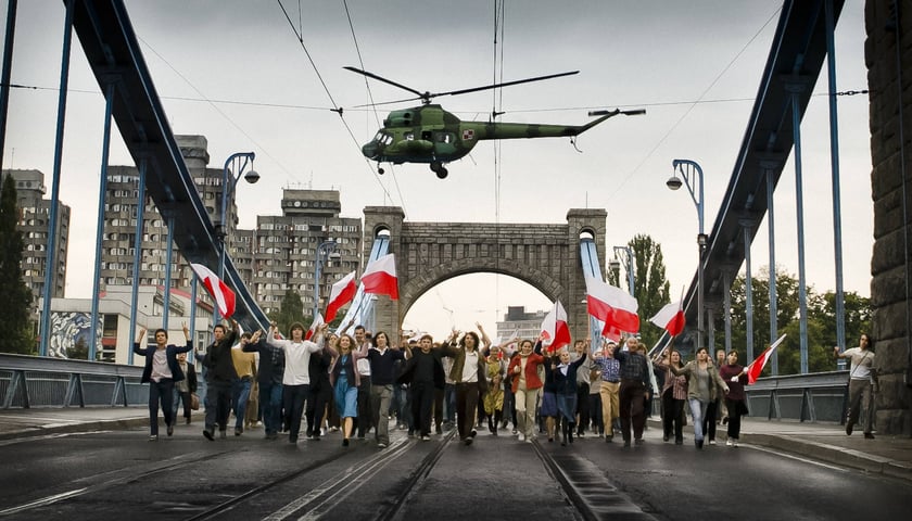 Śmigłowiec nad mostem, po którym idą ludzie z biało-czerwonymi flagami - scena z filmu „80 milionów” w reżyserii Waldemara Krzystka nakręcona we Wrocławiu na moście Grunwaldzkim