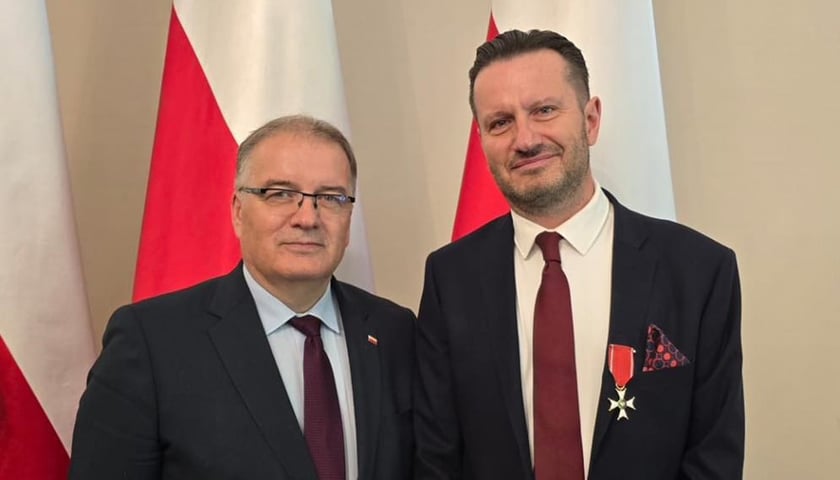 Prof. Krzysztof Kałwak (z prawej) i Andrzej Dera z Kancelarii Prezydenta RP. Na zdjęciu dwóch mężczyzn w ciemnych garniturach na tle biało-czerwonych flag