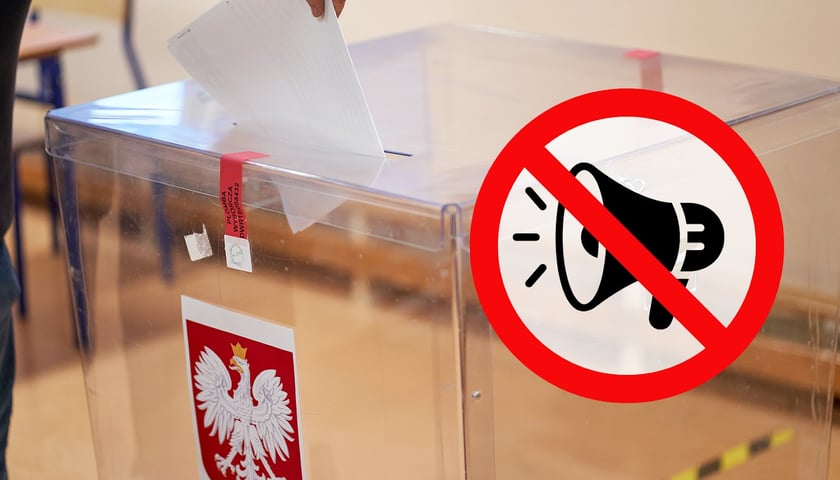 Na tablicy w czerwonym owalu zakazu przekreślony symbol głośnika / tuby, w tle urna w lokalu wyborczym